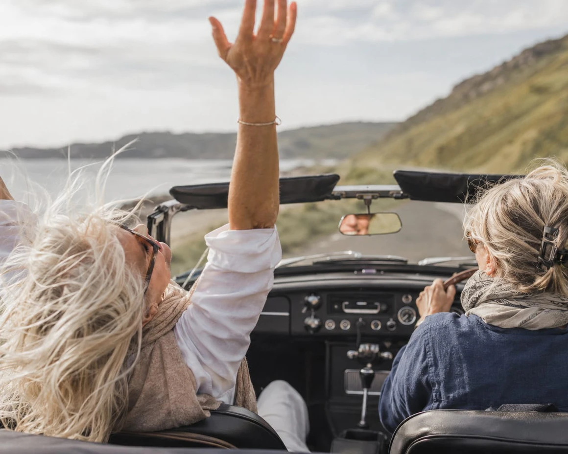 Two friends laughing in an open-top car on a coastal road, one with her arm in the air.