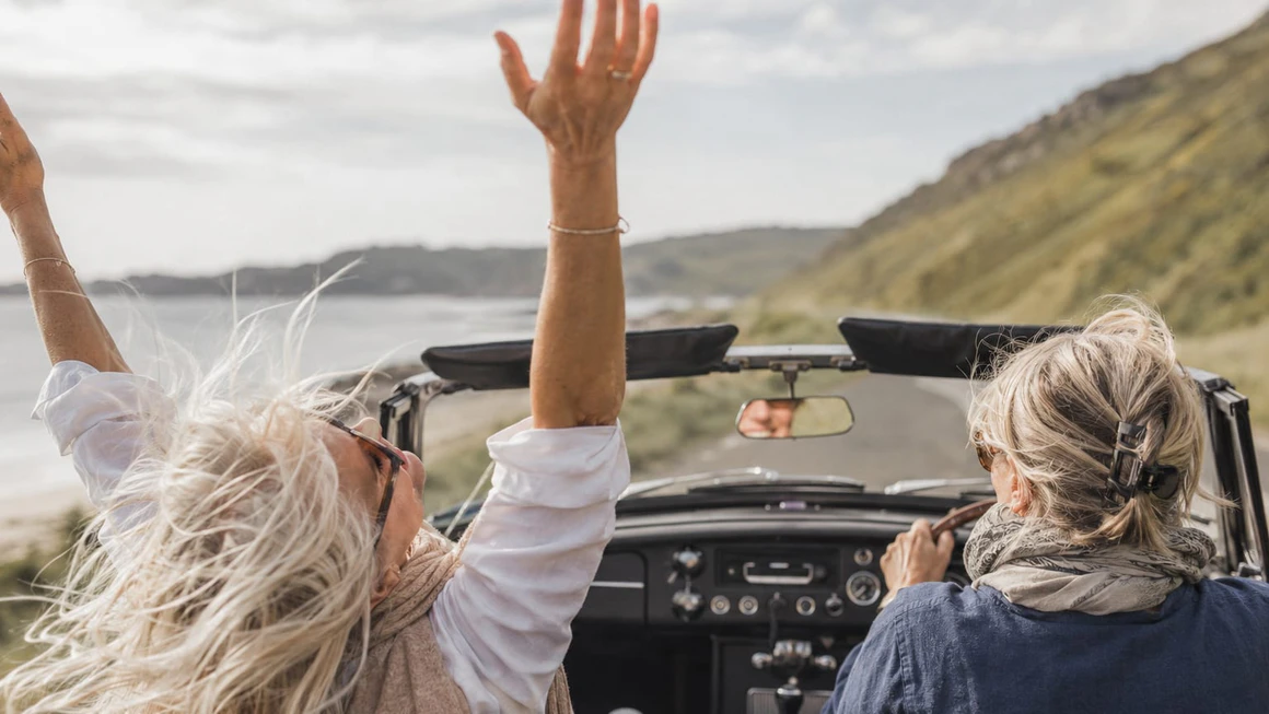 Two friends laughing in an open-top car on a coastal road, one with her arm in the air.