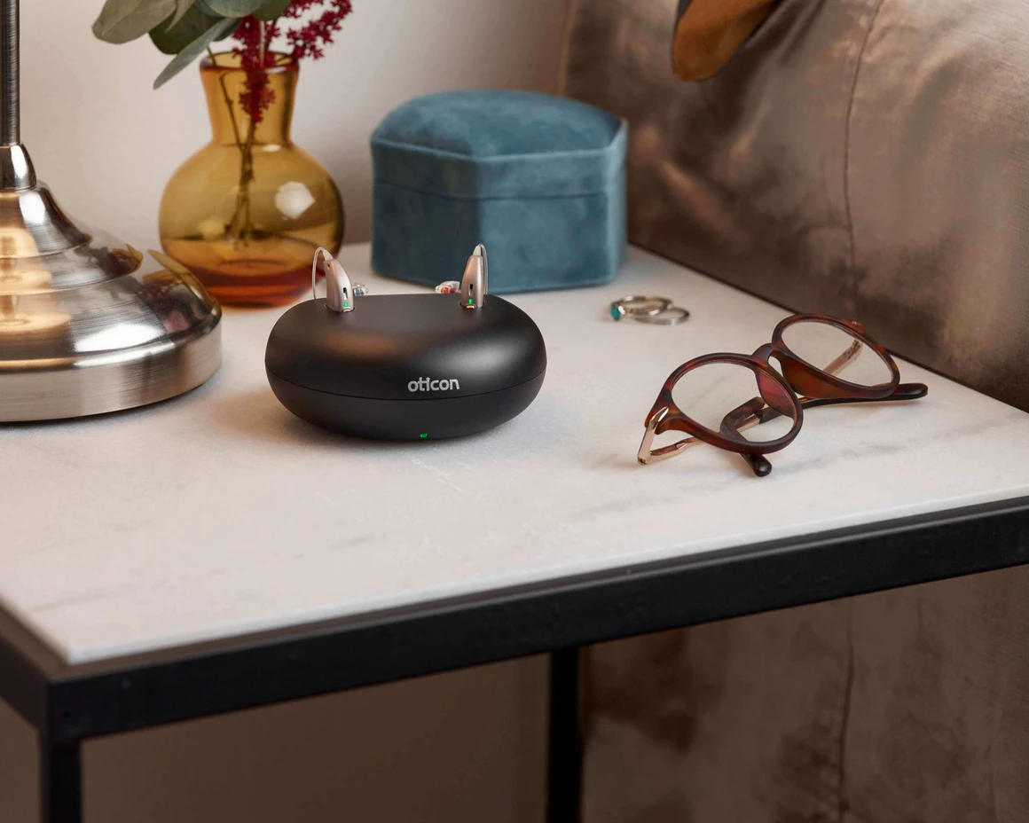 A pair of hearing aids Oticon Opn in a charging doc on a bedside table.