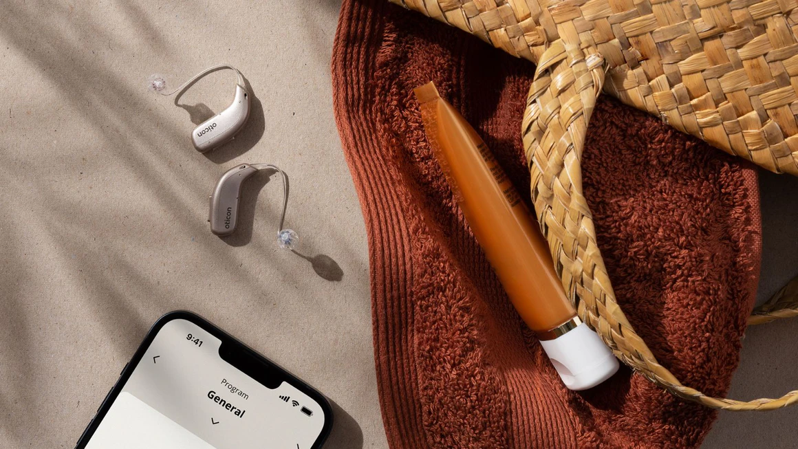 Oticon Real hearing aids and a phone with the Oticon app on beach sands.